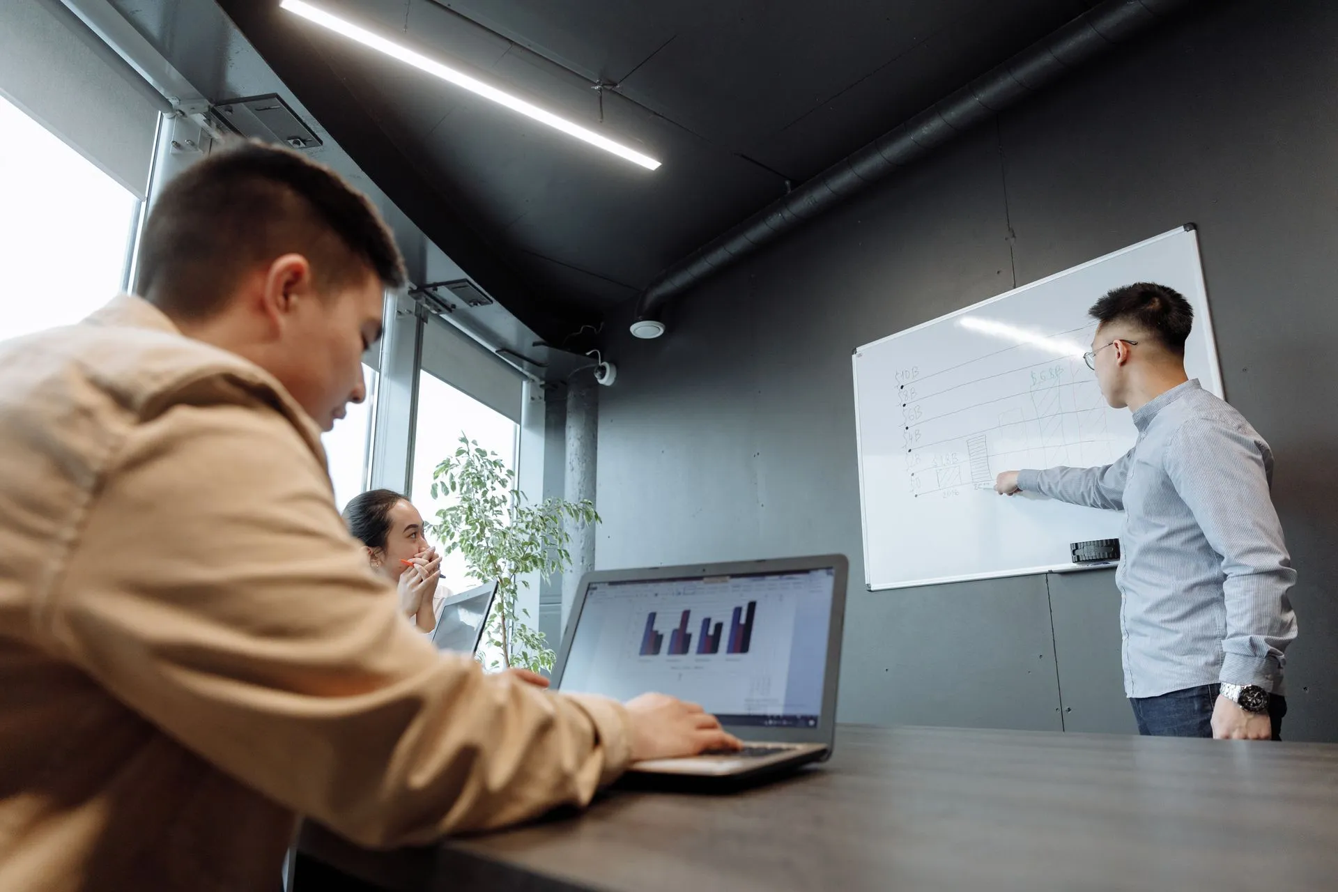 A colleague presenting at a whiteboard while a teammate follows on his laptop